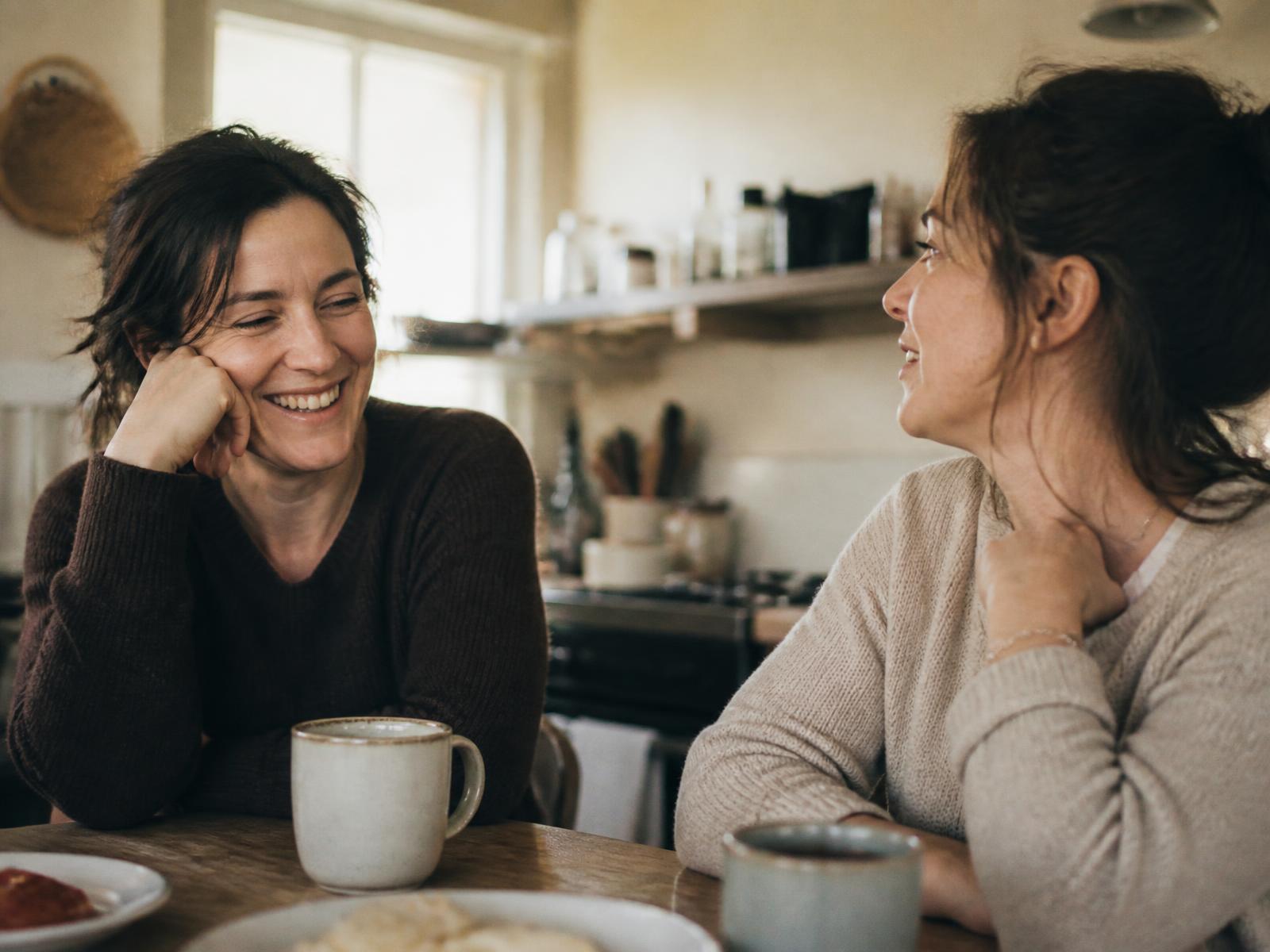 Two women standing together in a sunlit kitchen, talking and laughing during an ordinary everyday moment